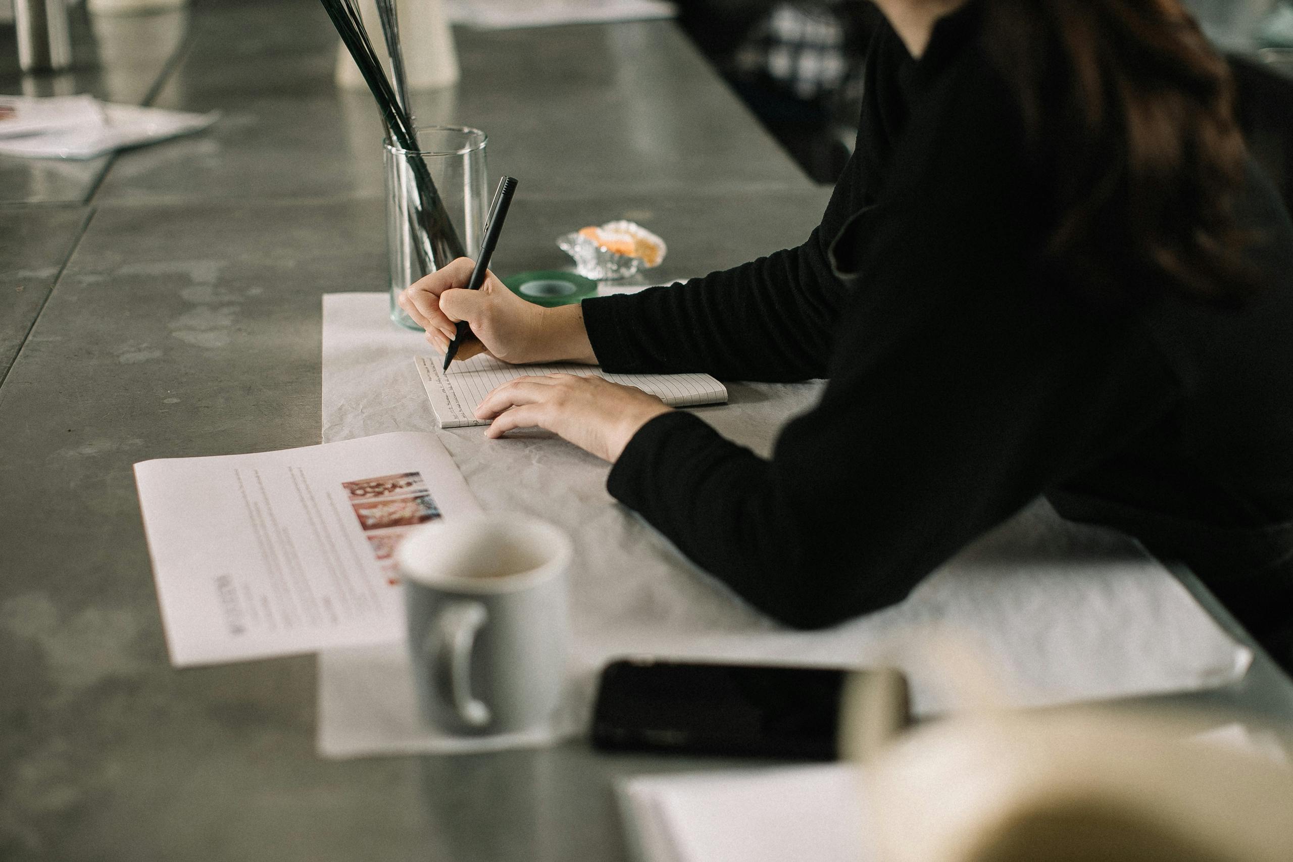 An adult taking notes at a desk in an office, focusing on work with coffee and documents nearby.