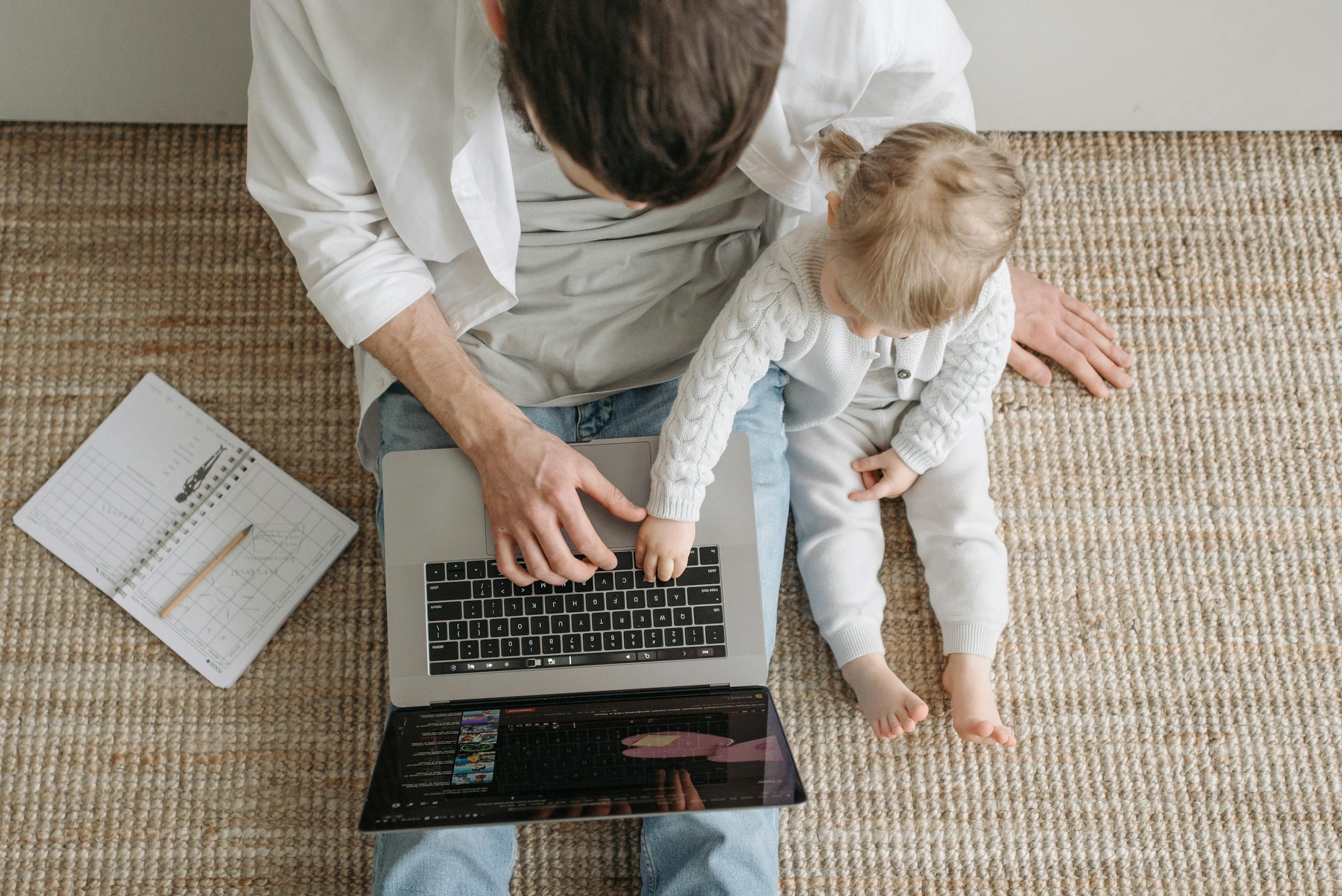 A father and baby sitting on a carpeted floor, interacting with a laptop in a cozy setting.