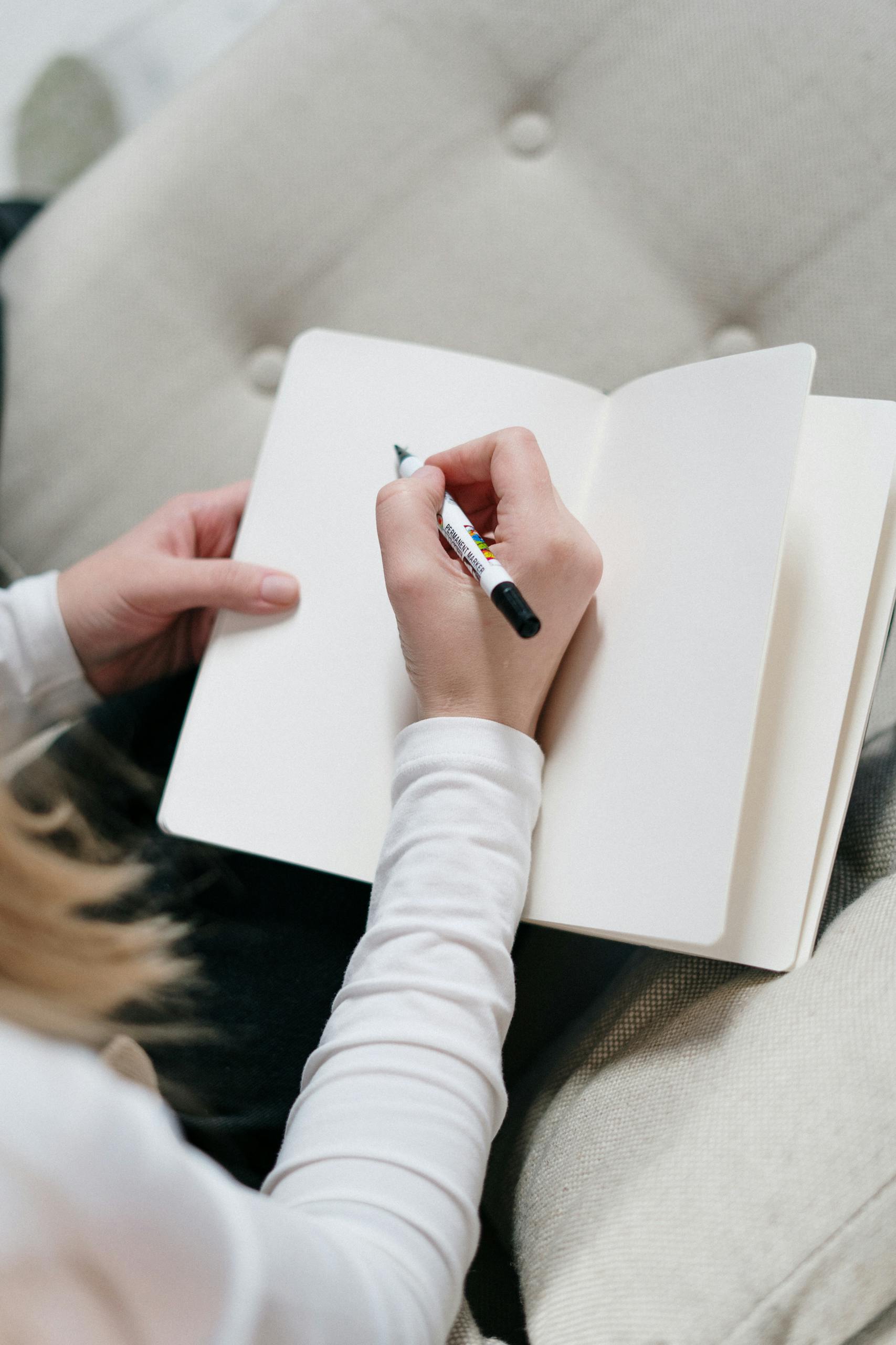 Top view of a woman writing in an open blank journal, sitting on a cozy chair indoors.
