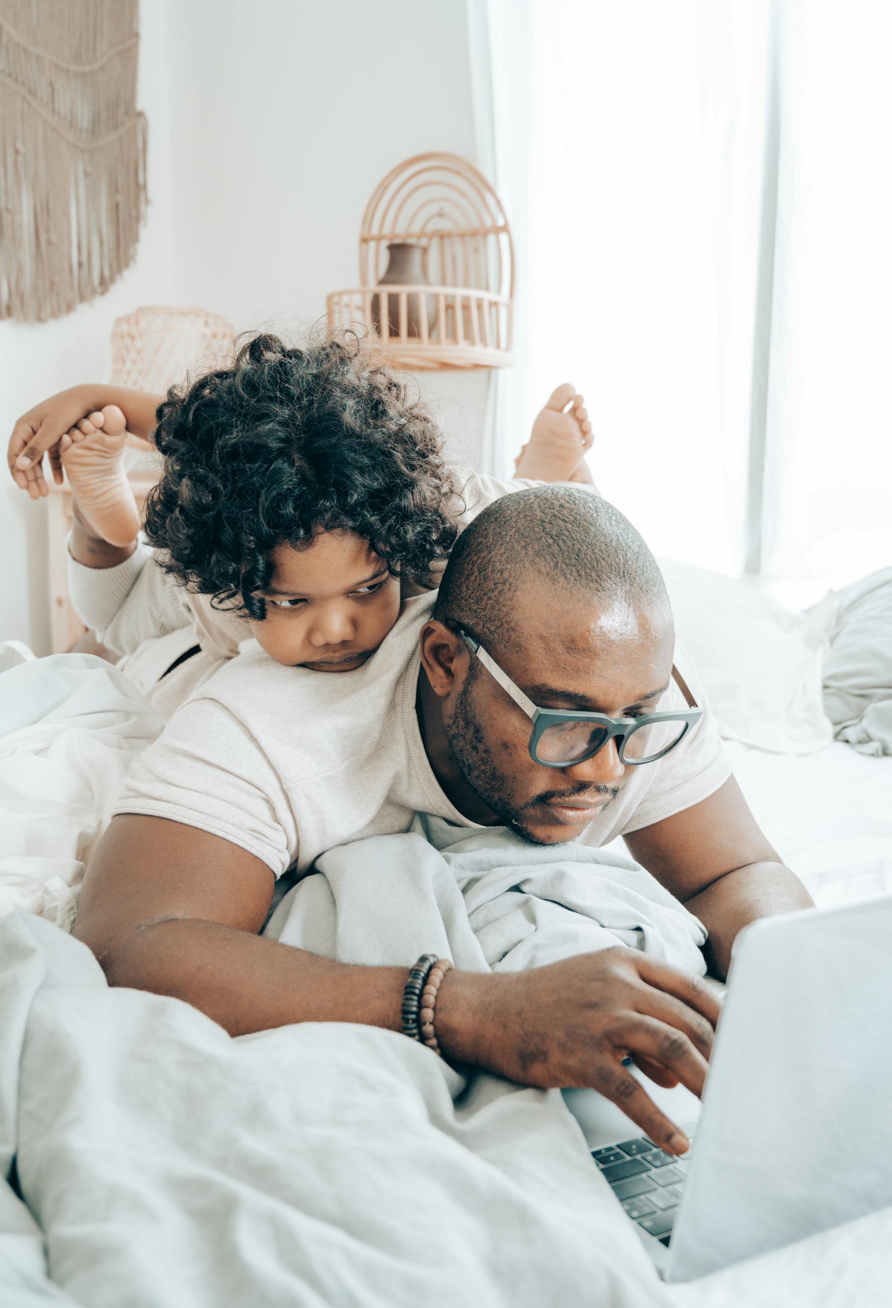 Young African American man typing on laptop while lying on bed with child on back in modern bedroom on weekend day